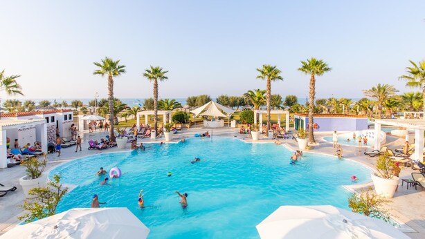 Una gran piscina al aire libre rodeada de palmeras y sillas de descanso, con vista al mar de fondo.