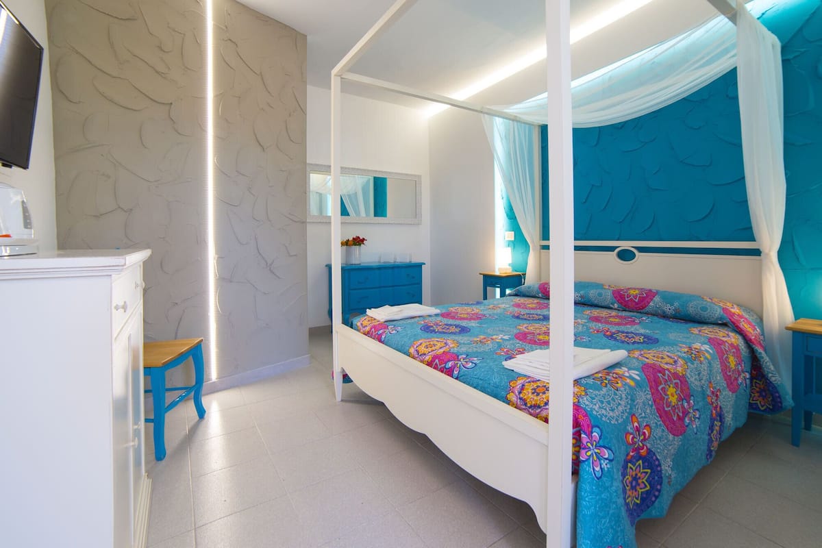A four-poster bed with a colorful floral bedspread, a white bedside table, a blue nightstand, a white dresser, and a mirror on the wall.