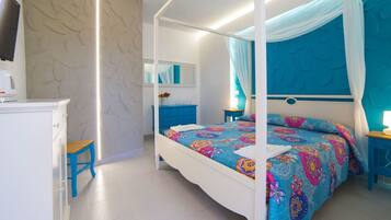 Un letto a baldacchino con una coperta colorata a motivi floreali, un comodino bianco, un tavolino da notte blu, una cassettiera bianca e uno specchio appeso al muro.