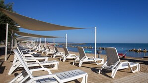 Un lounge frente a la playa con filas de sillas blancas bajo una pérgola, una terraza de madera y vistas al mar y afloramientos rocosos.