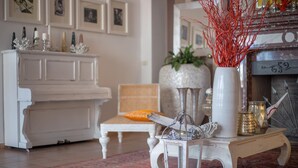 Una sala de estar con un piano blanco, una silla de madera, una mesa blanca con un farol y un jarrón con ramas rojas.