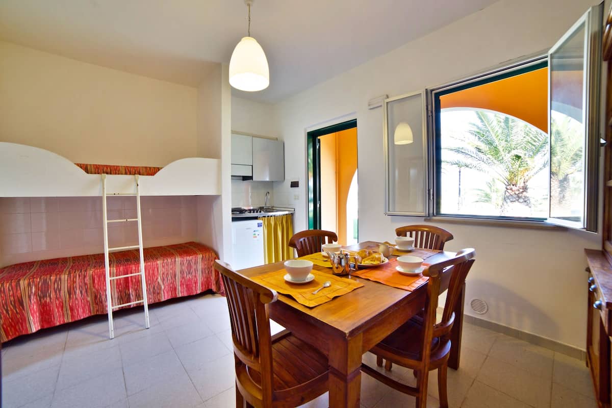 A dining area with a wooden table, chairs, and a bench. There is a window with a view of palm trees.