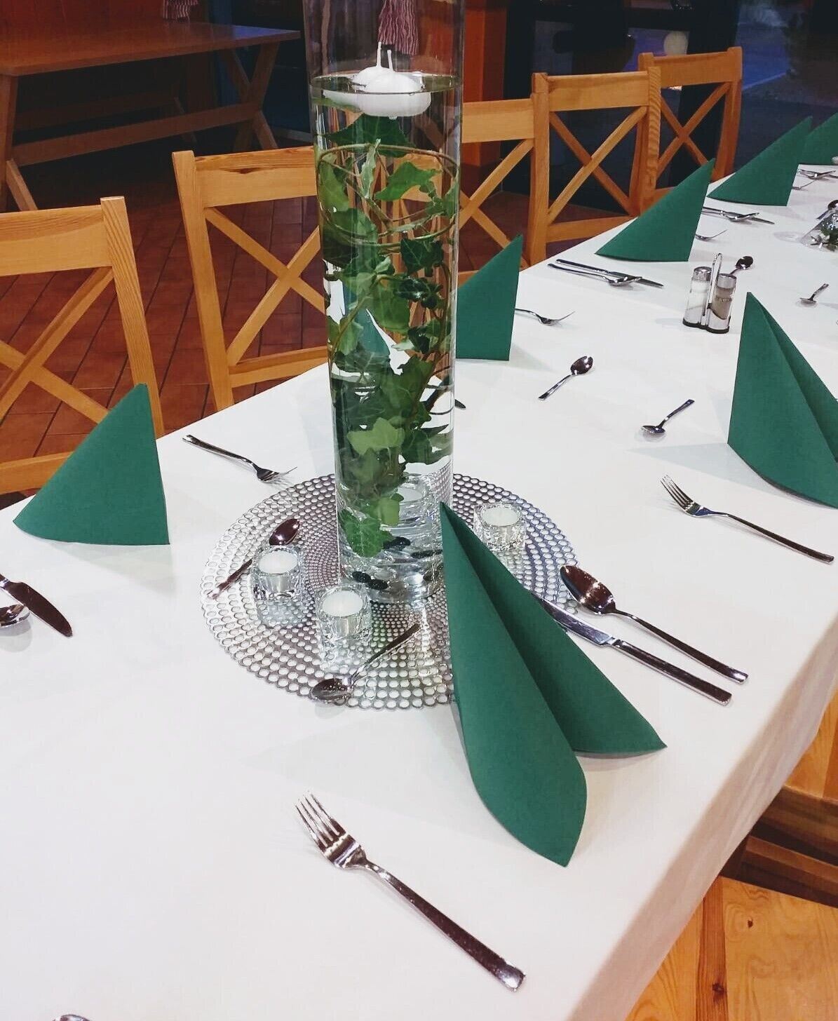 A table set for a meal with a decorative glass centerpiece containing green foliage and a candle.