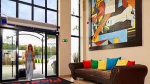 A hotel lobby with a large window, a reception desk, a brown leather sofa with colorful cushions, and a modern abstract painting on the wall.