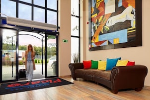 Un vestíbulo de hotel con un ventanal grande, un mostrador de recepción, un sofá de cuero marrón con cojines de colores y un cuadro abstracto moderno en la pared.