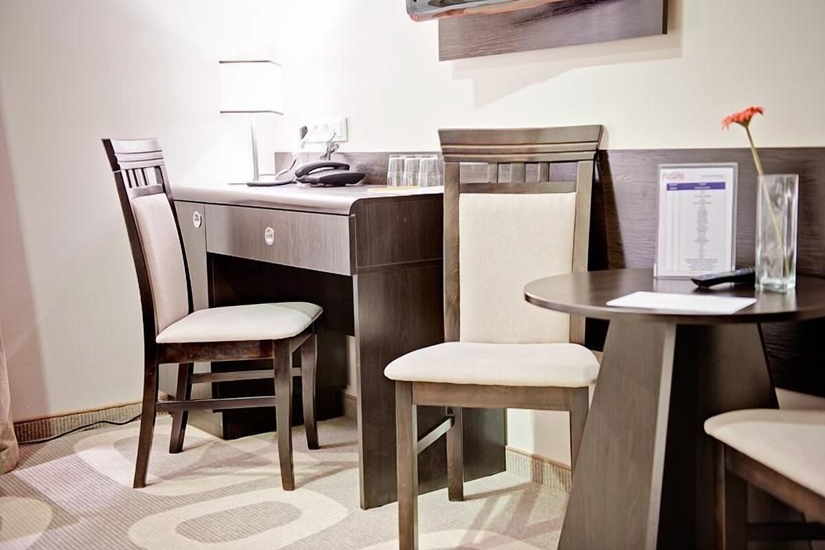 A hotel room with a desk, two chairs, a lamp, a vase with a flower, and a framed document on the wall.