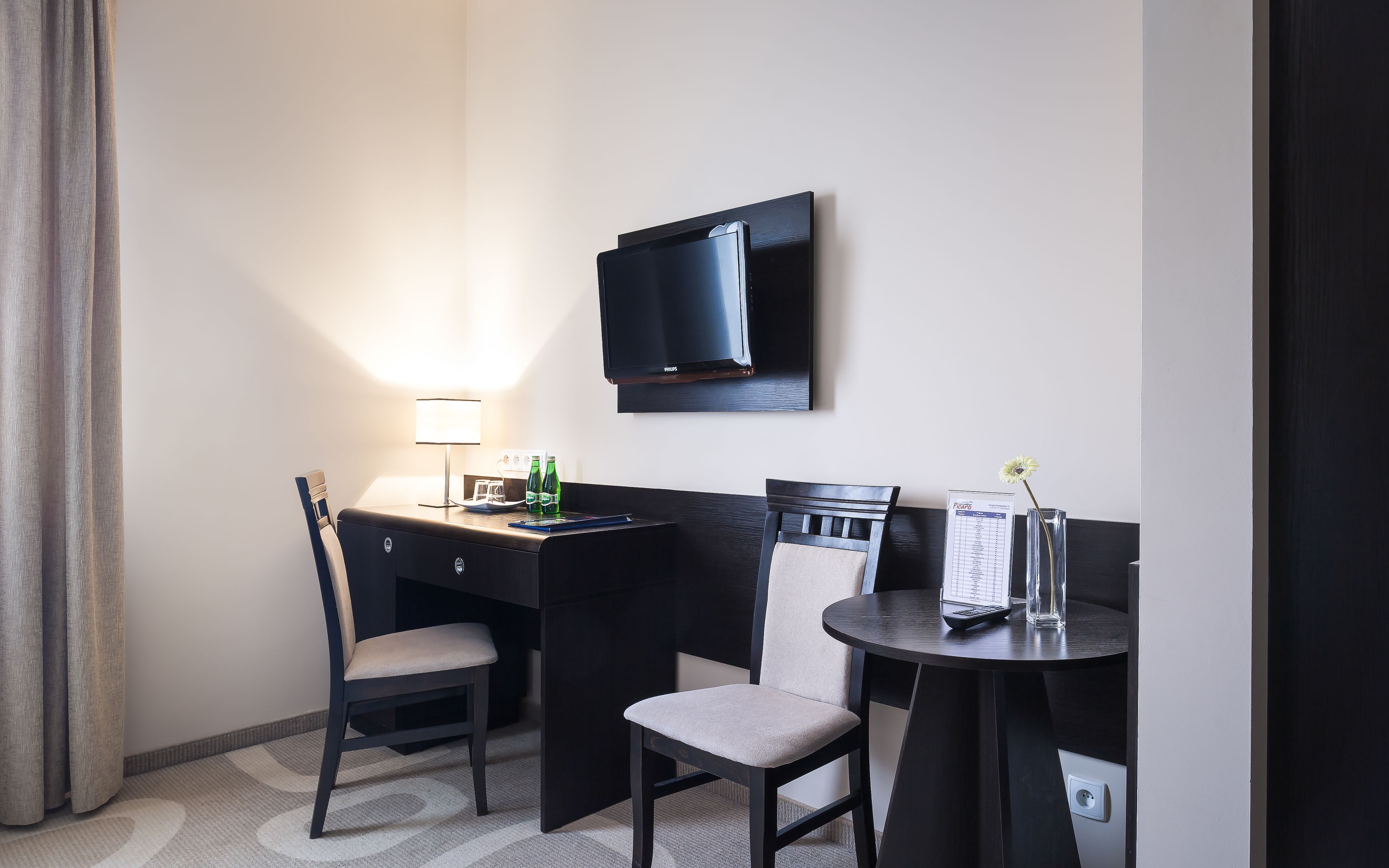 A hotel room with a desk, two chairs, a flat-screen TV, a lamp, a small table with a menu, and a vase with a flower.