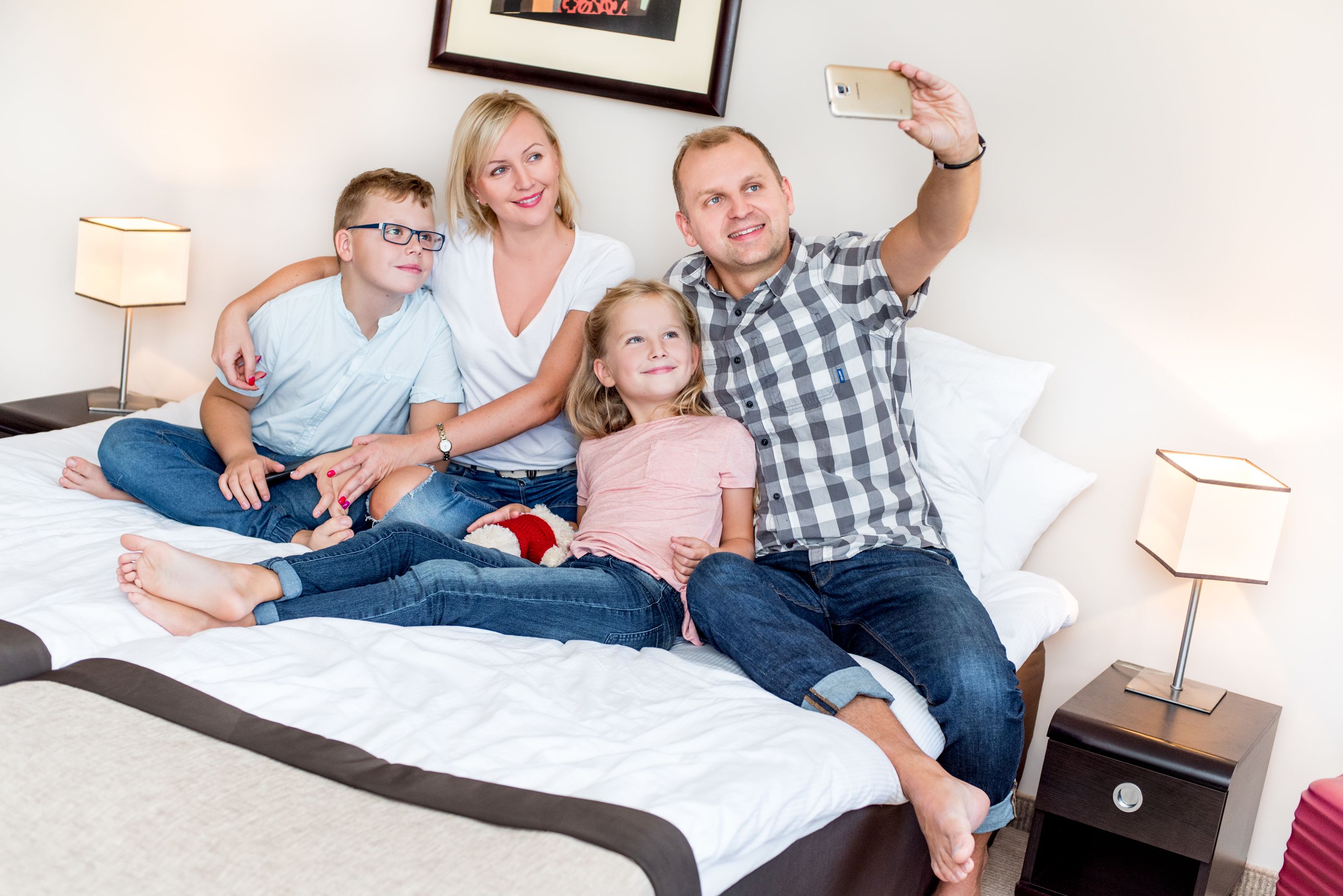 En familie på fire på sengen med et hvidt sengegavl, to sengeborde med lamper og et billede i ramme på væggen.