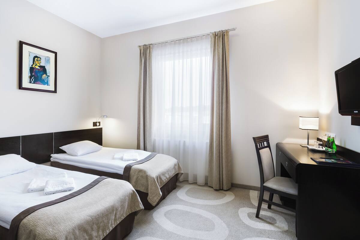 Habitación de hotel con dos camas, un escritorio con silla, televisor y ventana con cortinas.