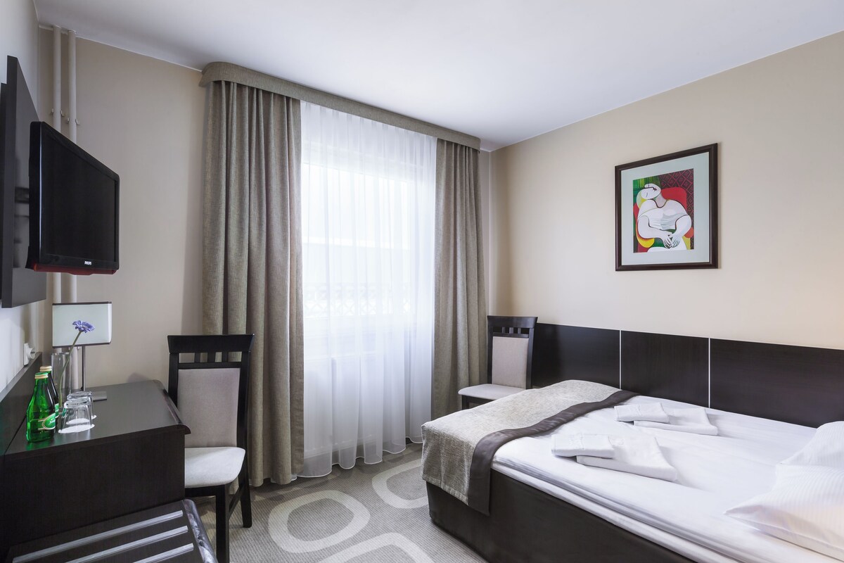 Ein Hotelzimmer mit Bett, Schreibtisch, Stuhl, Fernseher und einem Fenster mit Vorhängen.