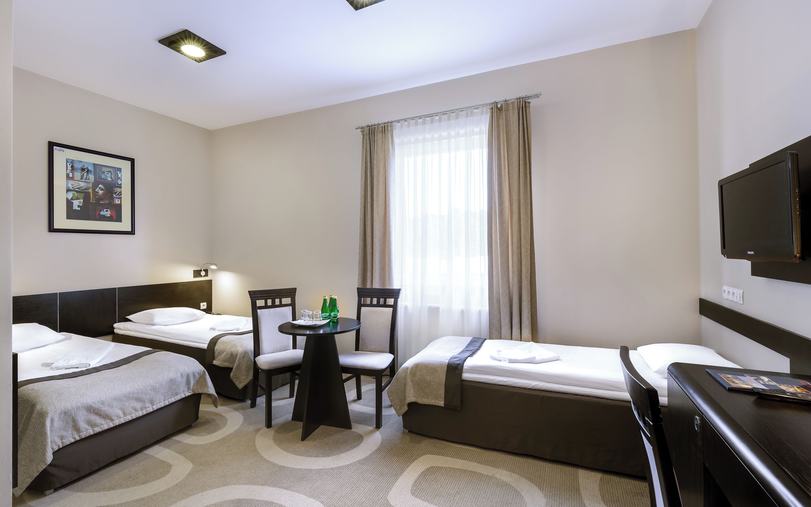 A hotel room with two beds, a desk with chairs, a television, and a window with curtains.