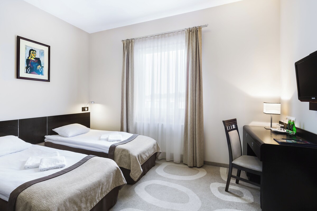 Ein Hotelzimmer mit zwei Betten, einem Schreibtisch mit Stuhl, einem Fernseher und einem Fenster mit Vorhängen.