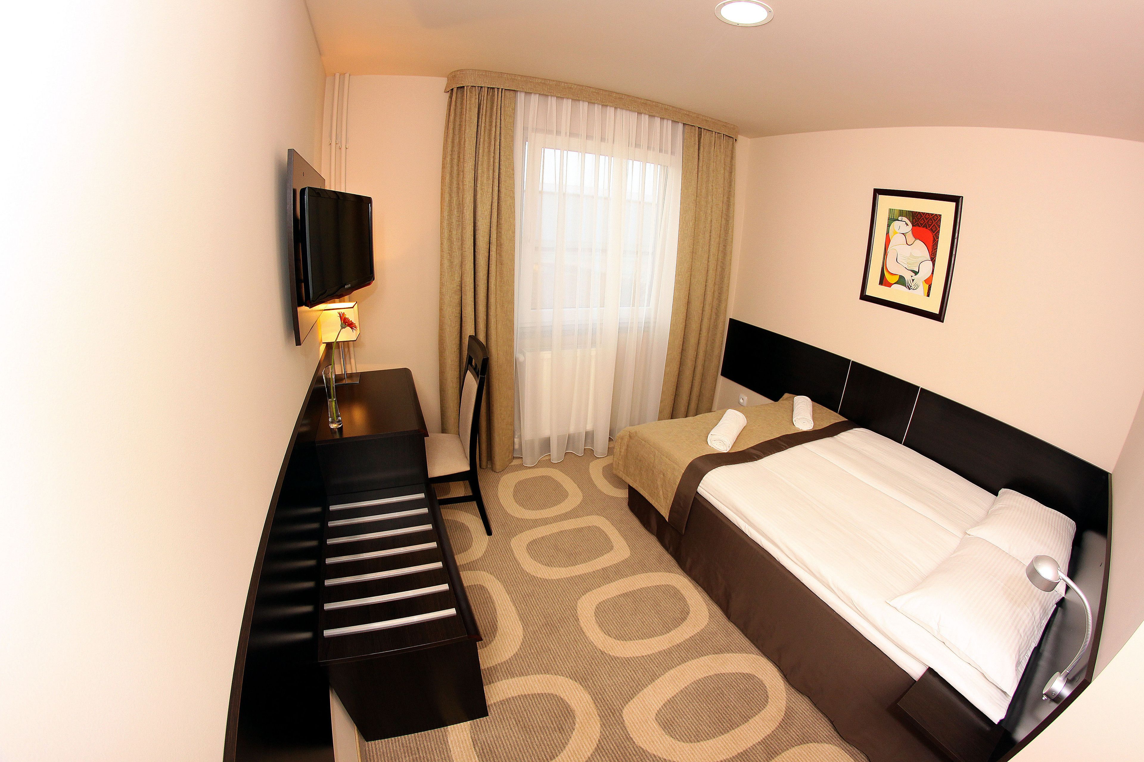 A hotel room with a bed, a desk with a chair, a TV, and a window with curtains.