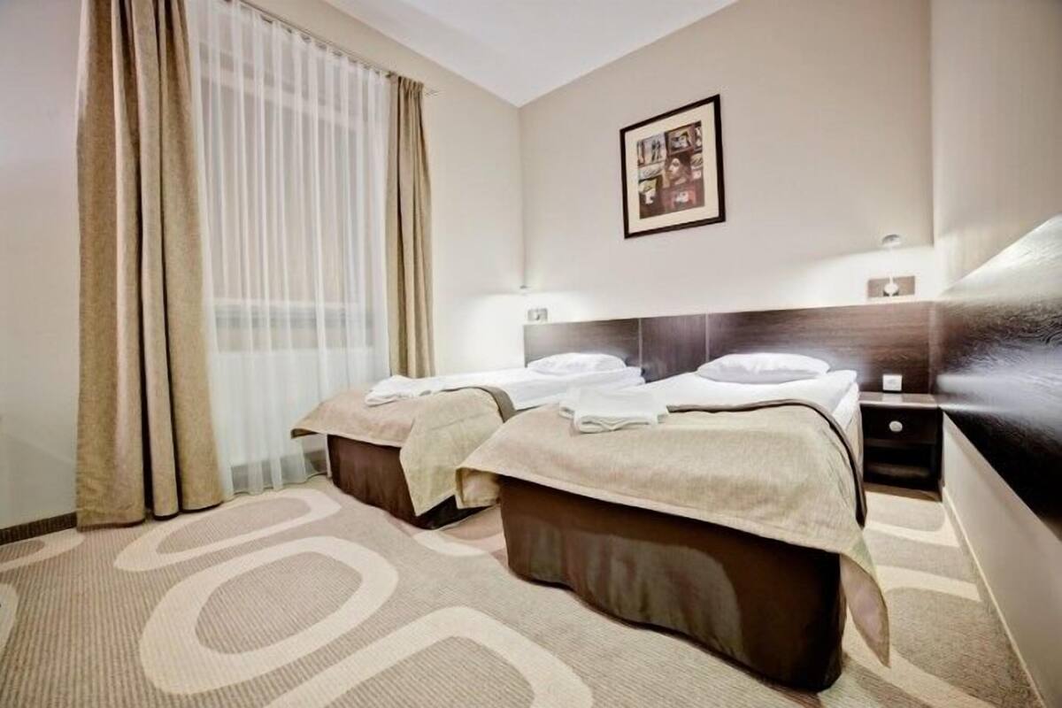 Habitación de hotel con dos camas, un ventanal con cortinas, cabecero, mesita de noche y un cuadro colgado en la pared.