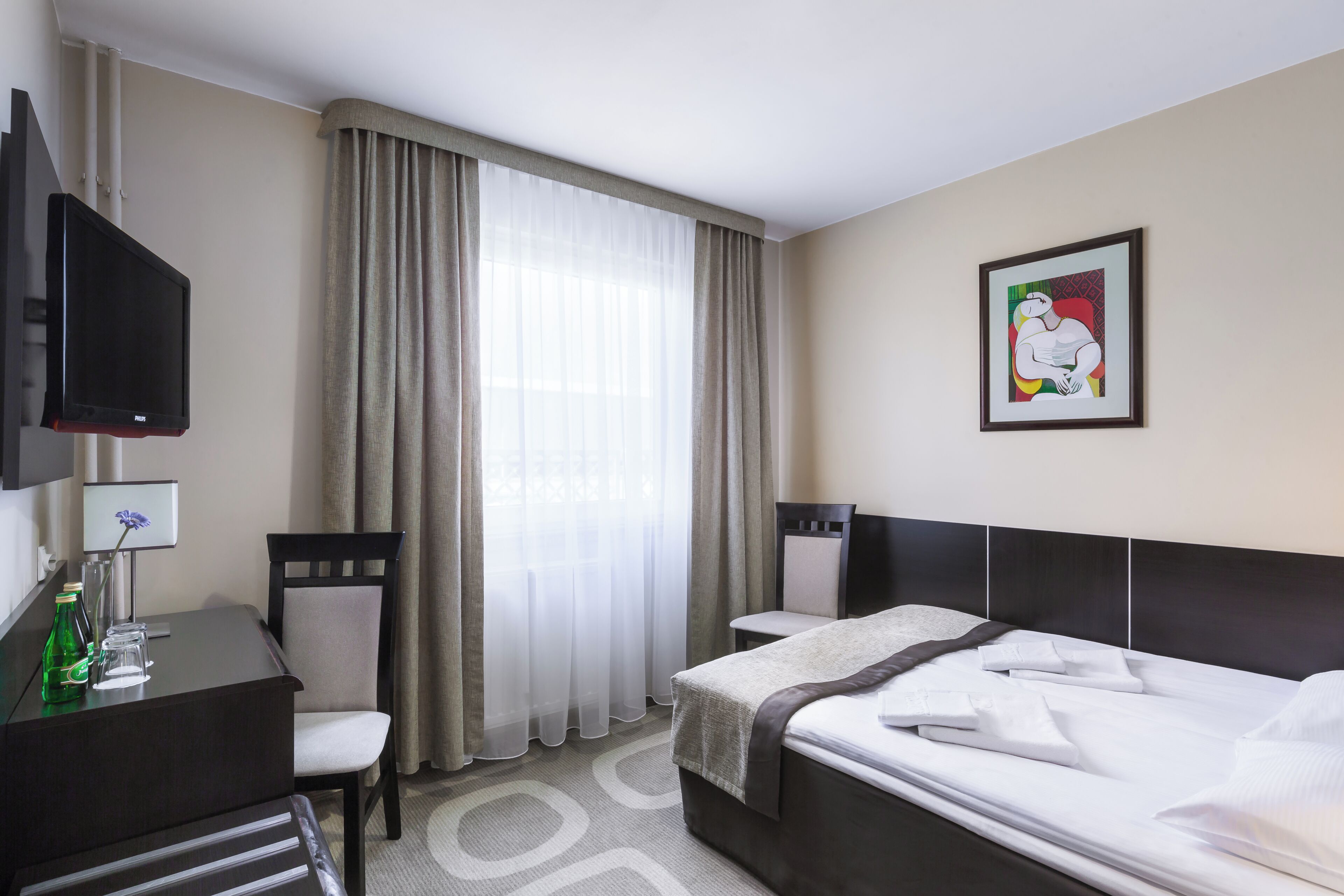 A hotel room with a bed, a desk with a chair, a television, a window with curtains, and a framed picture on the wall.