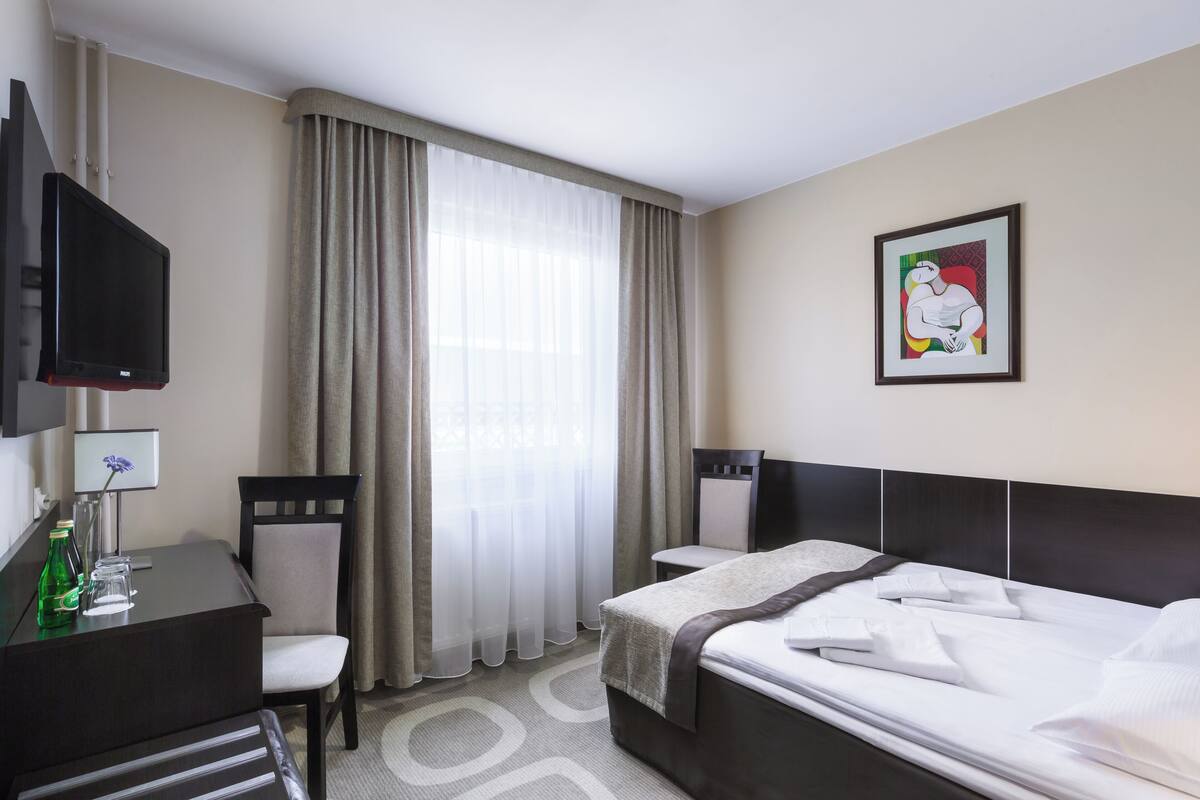 Una habitación de hotel con cama, escritorio y silla, televisión y ventana con cortinas.