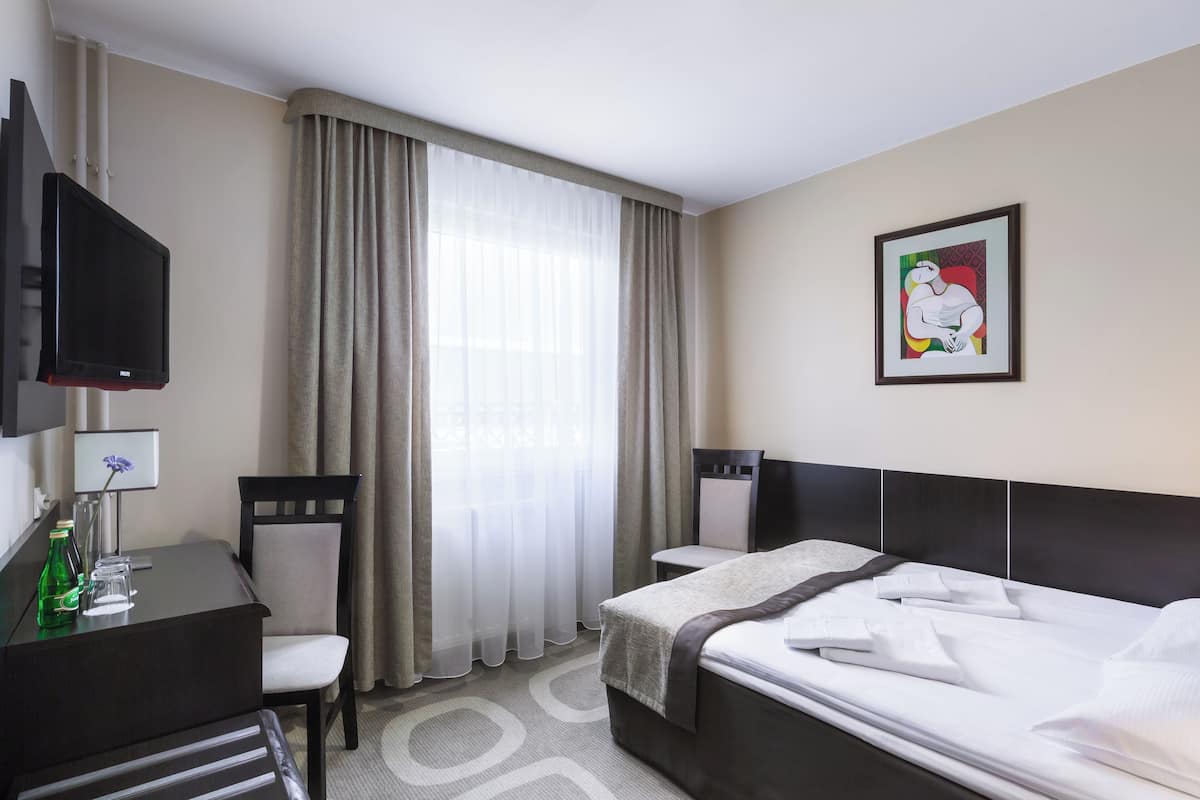 A hotel room with a bed, a desk with a chair, a television, a window with curtains, and a framed picture on the wall.