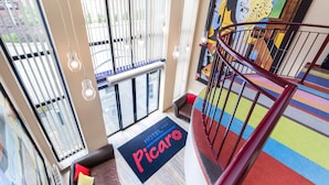 Un vestíbulo de hotel con una alfombra colorida, un felpudo negro con el texto 'HOTEL Picar', un sofá de cuero marrón y un cuadro abstracto grande.