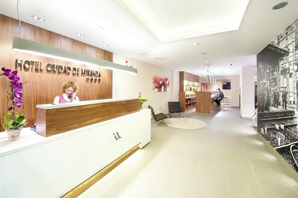 A modern hotel reception area with a wooden reception desk, a receptionist, and a large poster on the wall. - Hotel Sercotel Ciudad de Miranda (Miranda de Ebro)