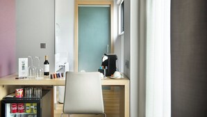 Habitación de hotel con un escritorio de madera, un refrigerador lleno de bebidas, una cafetera y vistas a través de una puerta.