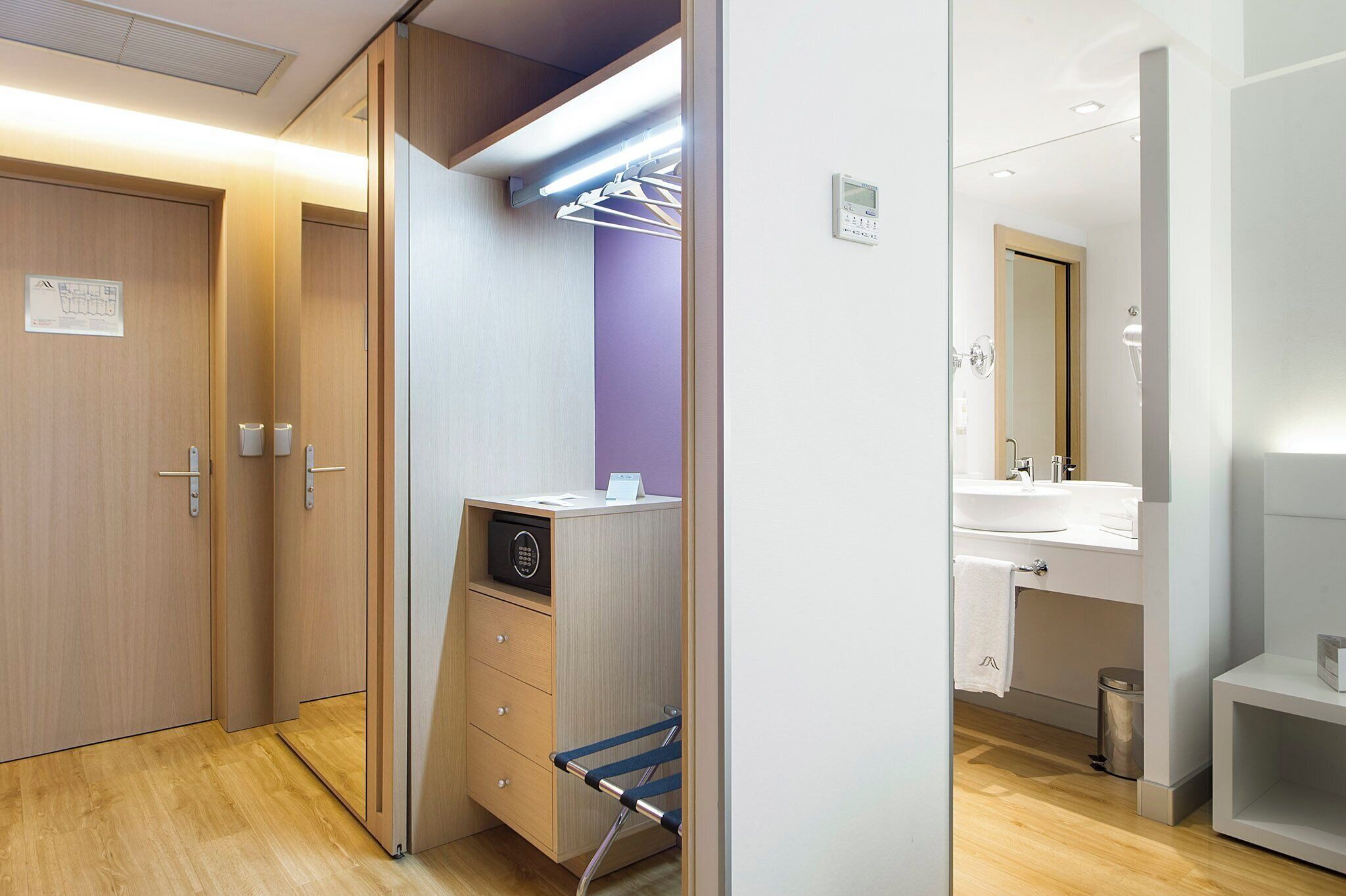 A modern hotel room with a wooden wardrobe, a built-in desk with a radio, a small seating area, and a bathroom with a sink and mirror.