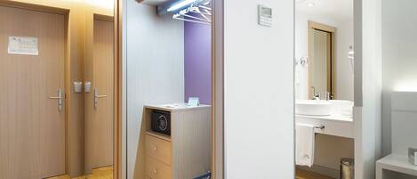 A modern hotel room with a wooden wardrobe, a built-in desk with a radio, a small seating area, and a bathroom with a sink and mirror.
