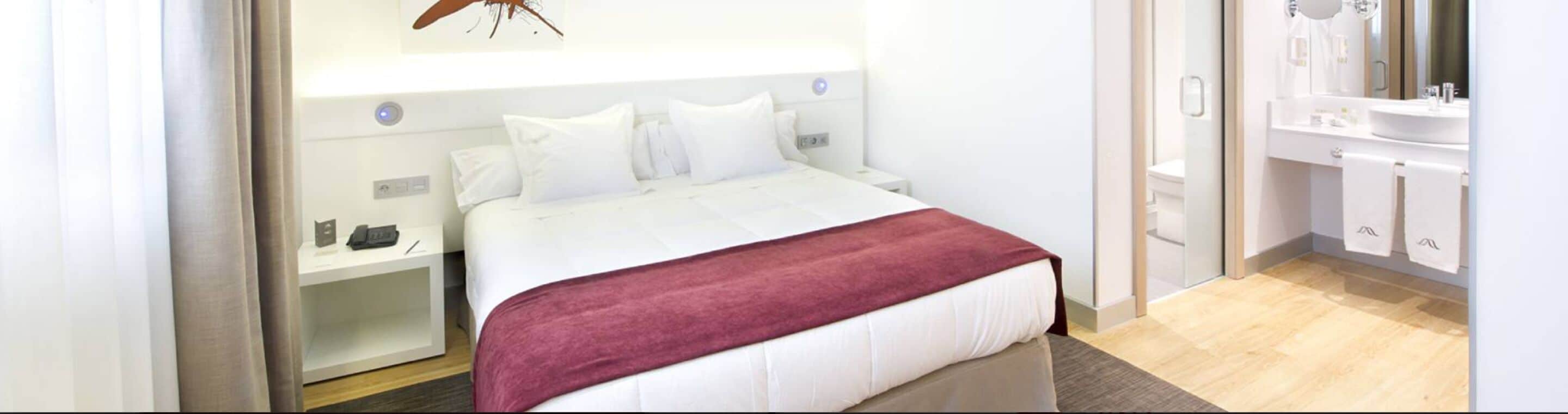 A hotel room with a bed, a bedside table with a phone and a pen, a red throw blanket, and a bathroom with a sink and mirror.