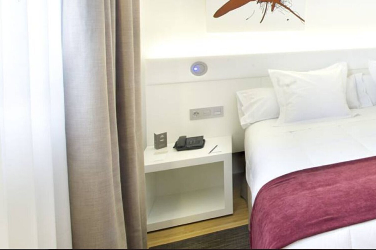 A hotel room with a bed, a bedside table with a phone and a pen, a red throw blanket, and a bathroom with a sink and mirror.
