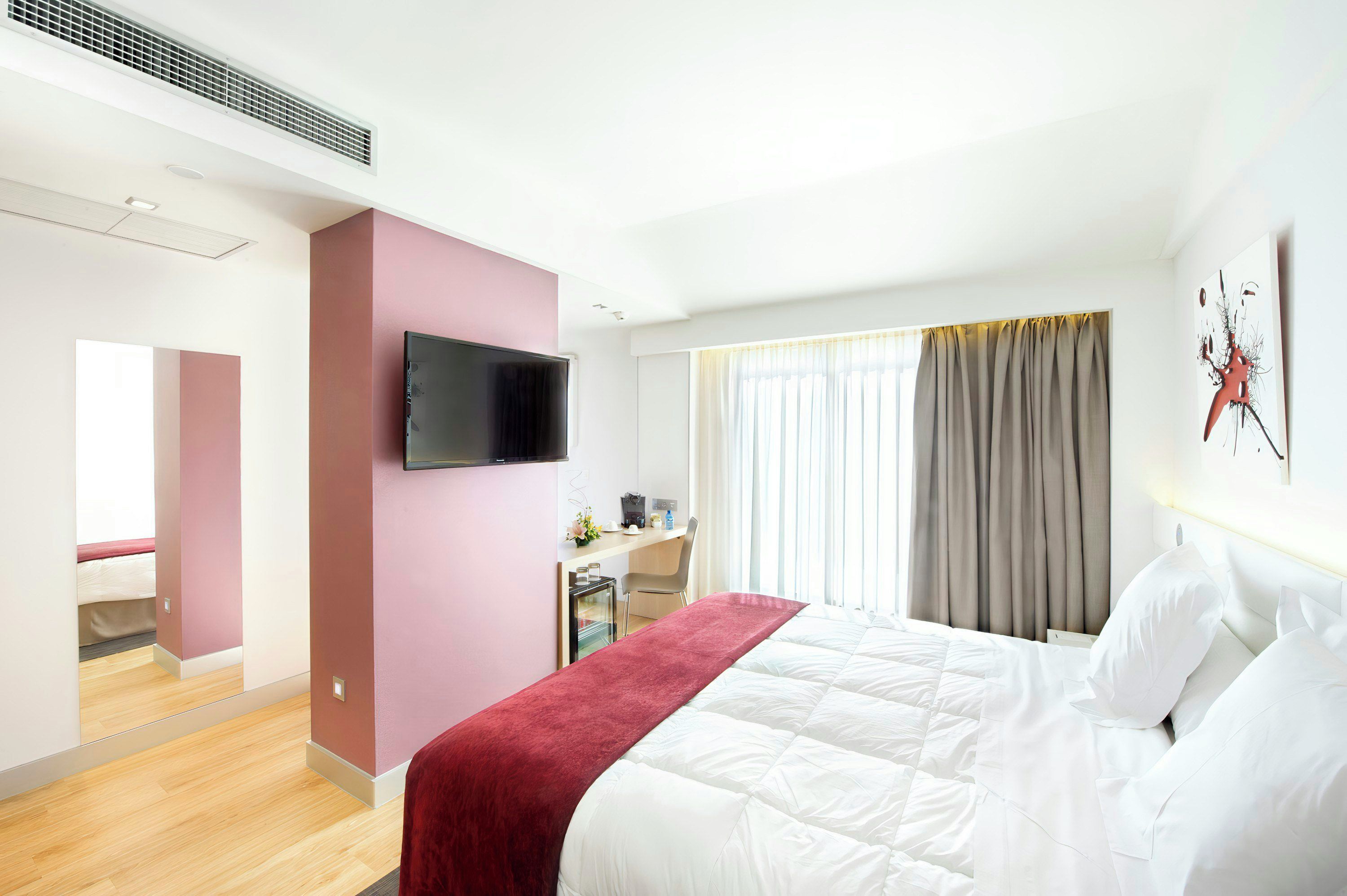 Habitación de hotel con cama, televisión, escritorio con silla, ventana con cortinas y una obra de arte colgada en la pared.