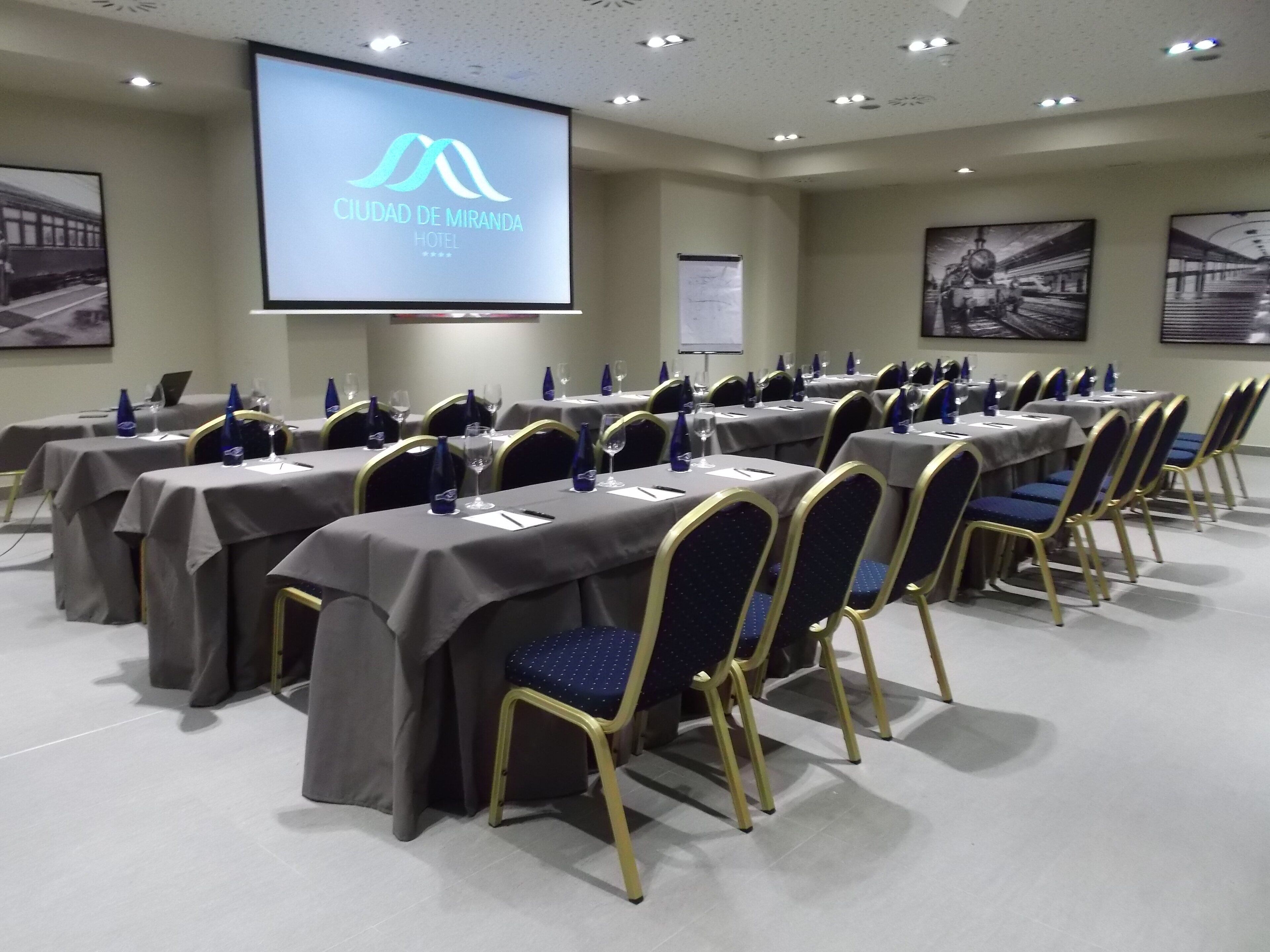 Une salle de conférence avec des tables disposées pour une réunion, un écran de projection affichant le nom de l’hôtel et des tableaux encadrés accrochés aux murs.