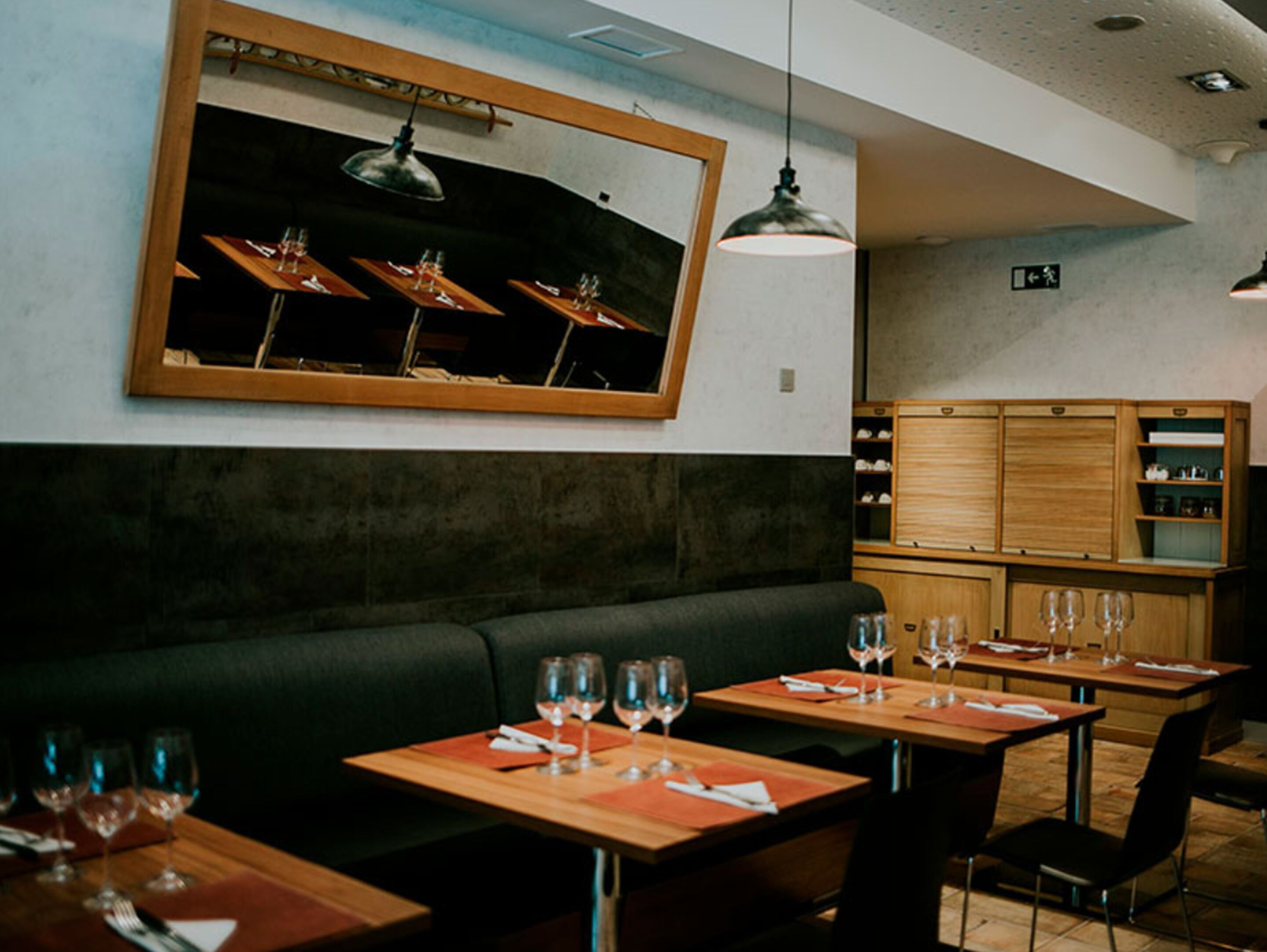 A restaurant interior with wooden tables, black cushioned seating, and pendant lighting.