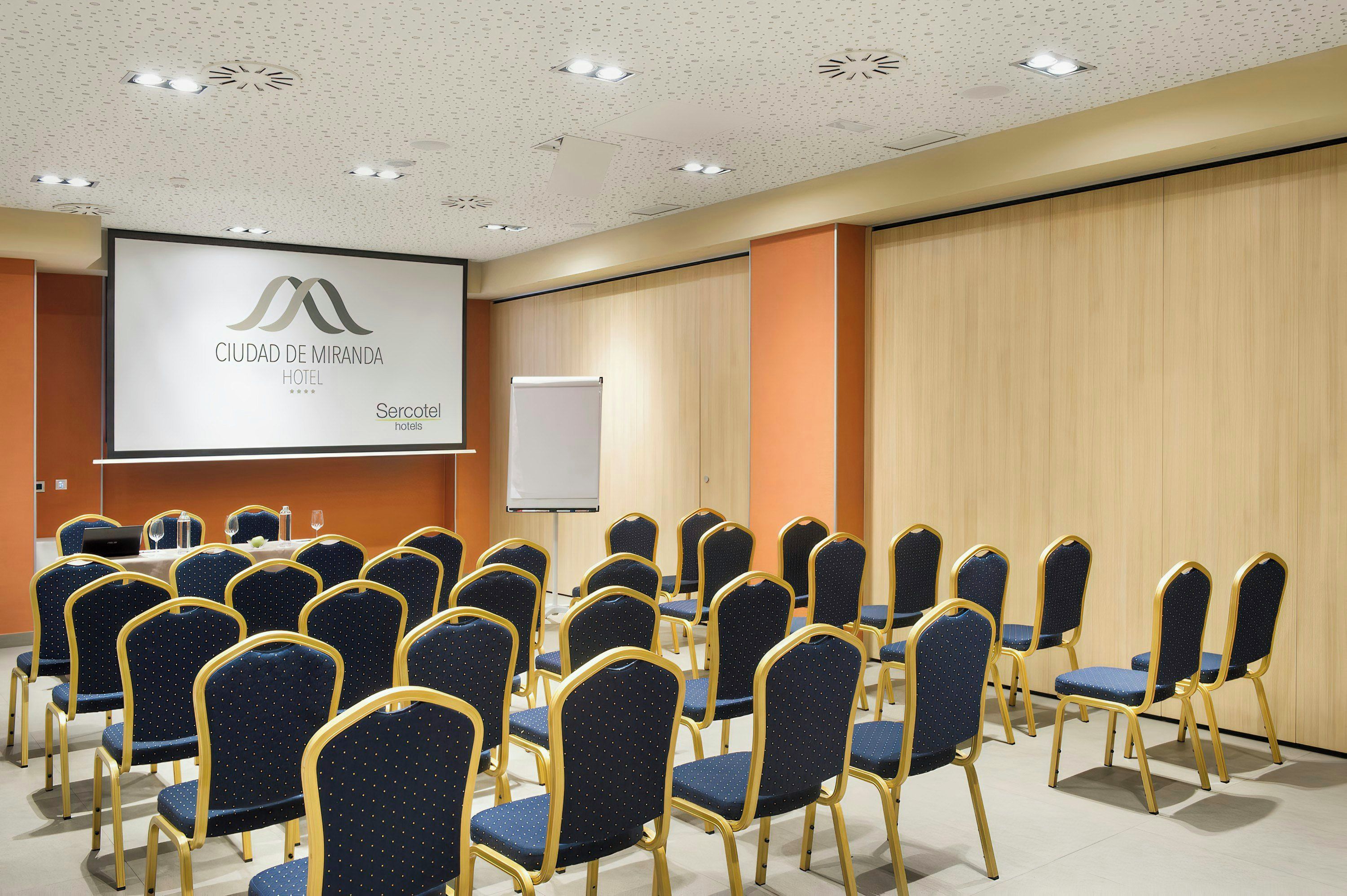 Un salón de conferencias con filas de sillas, una pantalla de proyección que muestra 'CIUDAD DE MIRANDA HOTEL' y 'Sercotel hotels', y una pizarra.