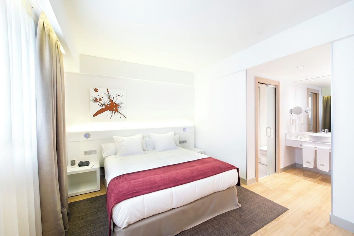 A modern hotel room with a large bed, bedside tables, a nightstand, a red throw blanket, a wall art piece, a bathroom with a mirror and sink.