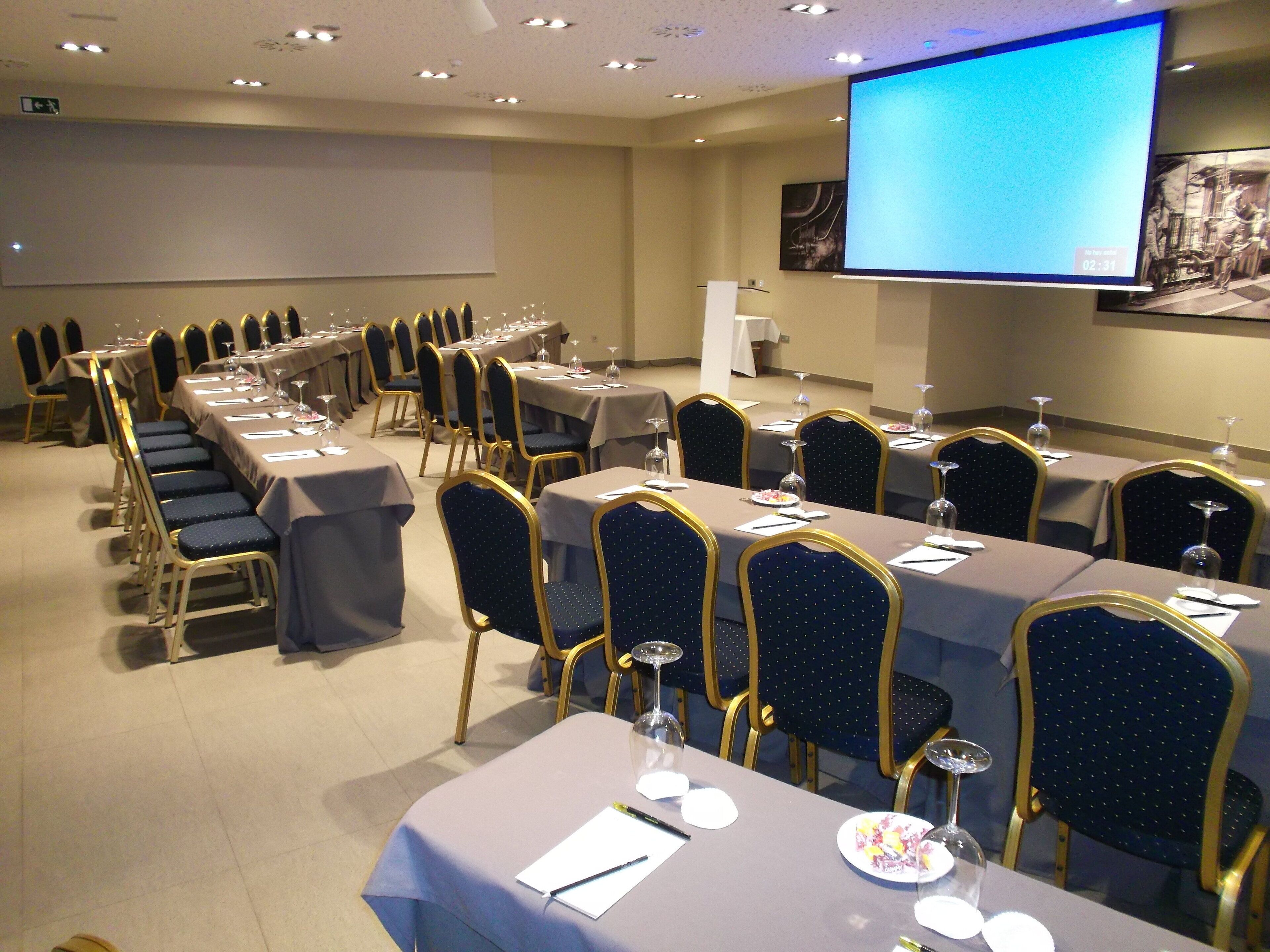 Un salón de conferencias con filas de mesas y sillas, equipado con tarjetas de identificación, vasos de agua y una pantalla de proyector mostrando una presentación.