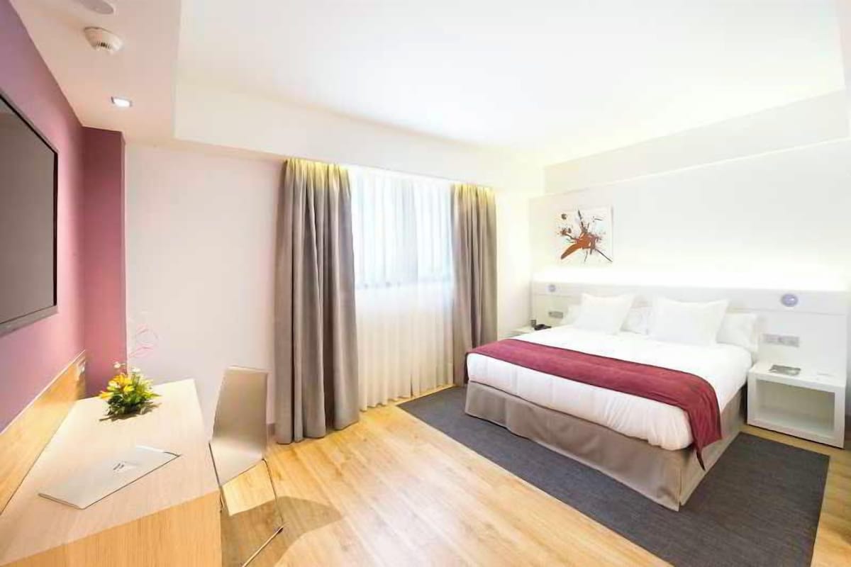 A hotel room with a bed, bedside tables, a desk with a chair, a TV, and a window with curtains.