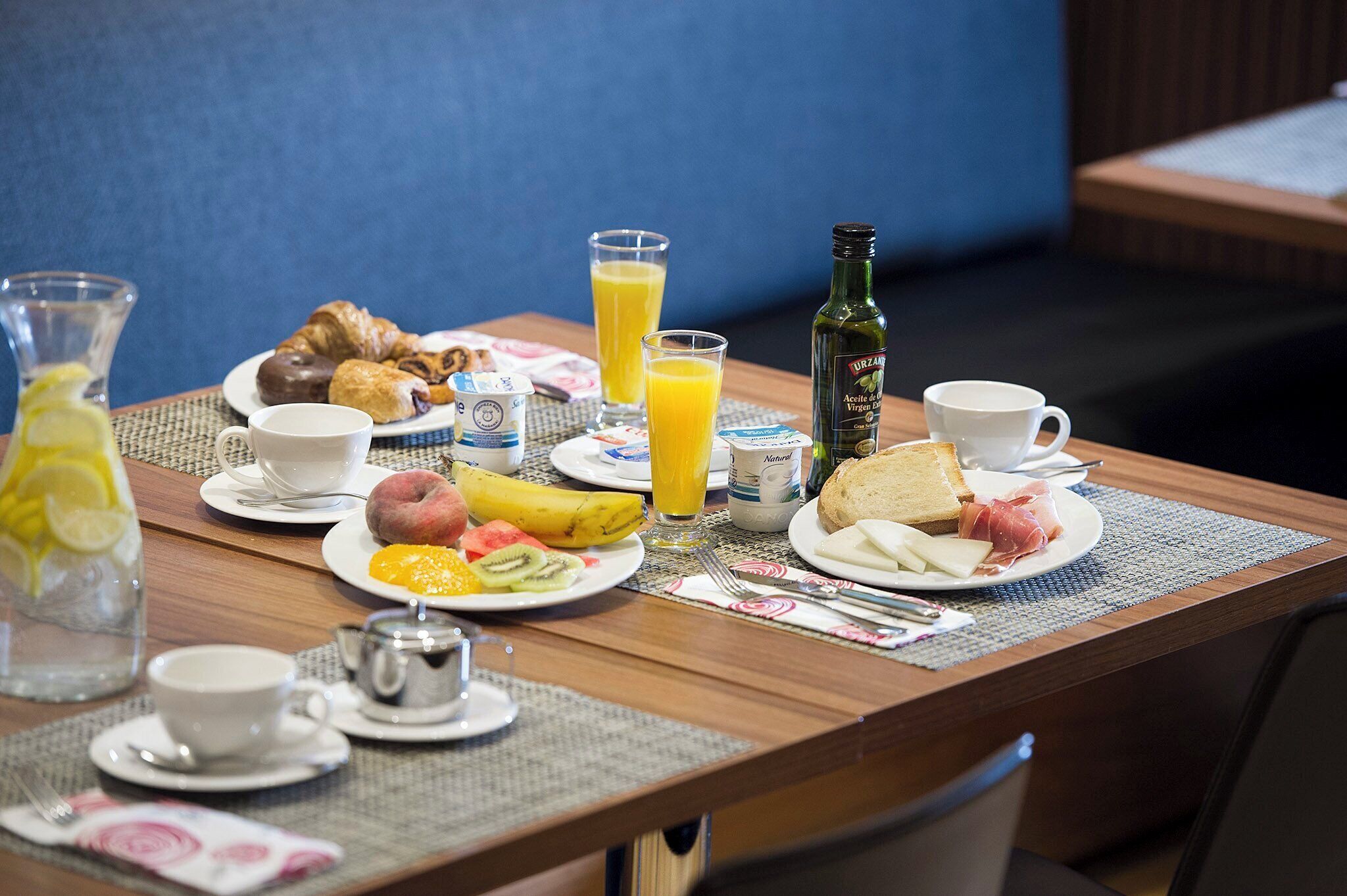 Une table de petit-déjeuner avec des croissants, des fruits, du jus et une bouteille d’huile d’olive.