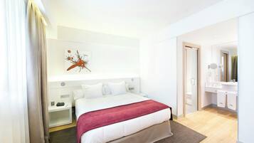 A hotel room with a bed, bedside tables, a nightstand, a wall-mounted artwork, a bathroom with a sink and mirror, and a wooden floor.