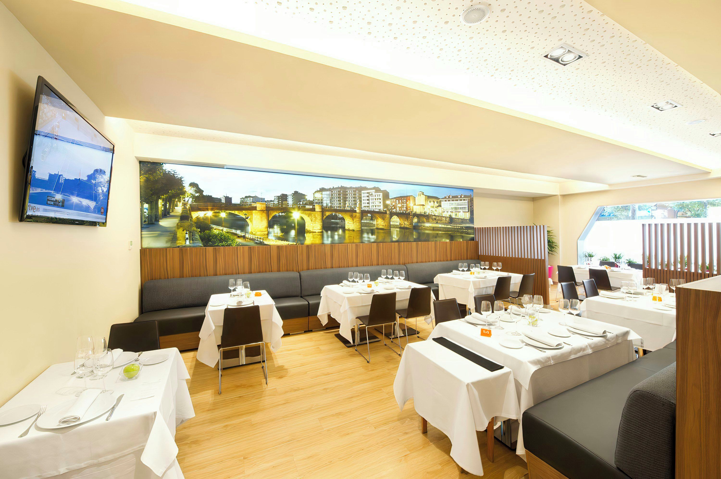A restaurant with multiple tables, a large cityscape painting on the wall, and a television screen displaying a sports event.