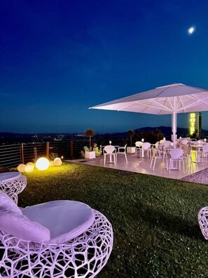 An outdoor seating area with round white wicker chairs and a large umbrella, set against a night sky with a visible moon.
