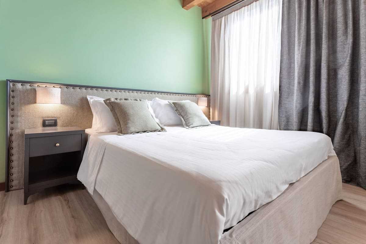 Una cama bien tendida con sábanas blancas y dos almohadas, una mesita de noche con una lámpara y una ventana con cortinas.