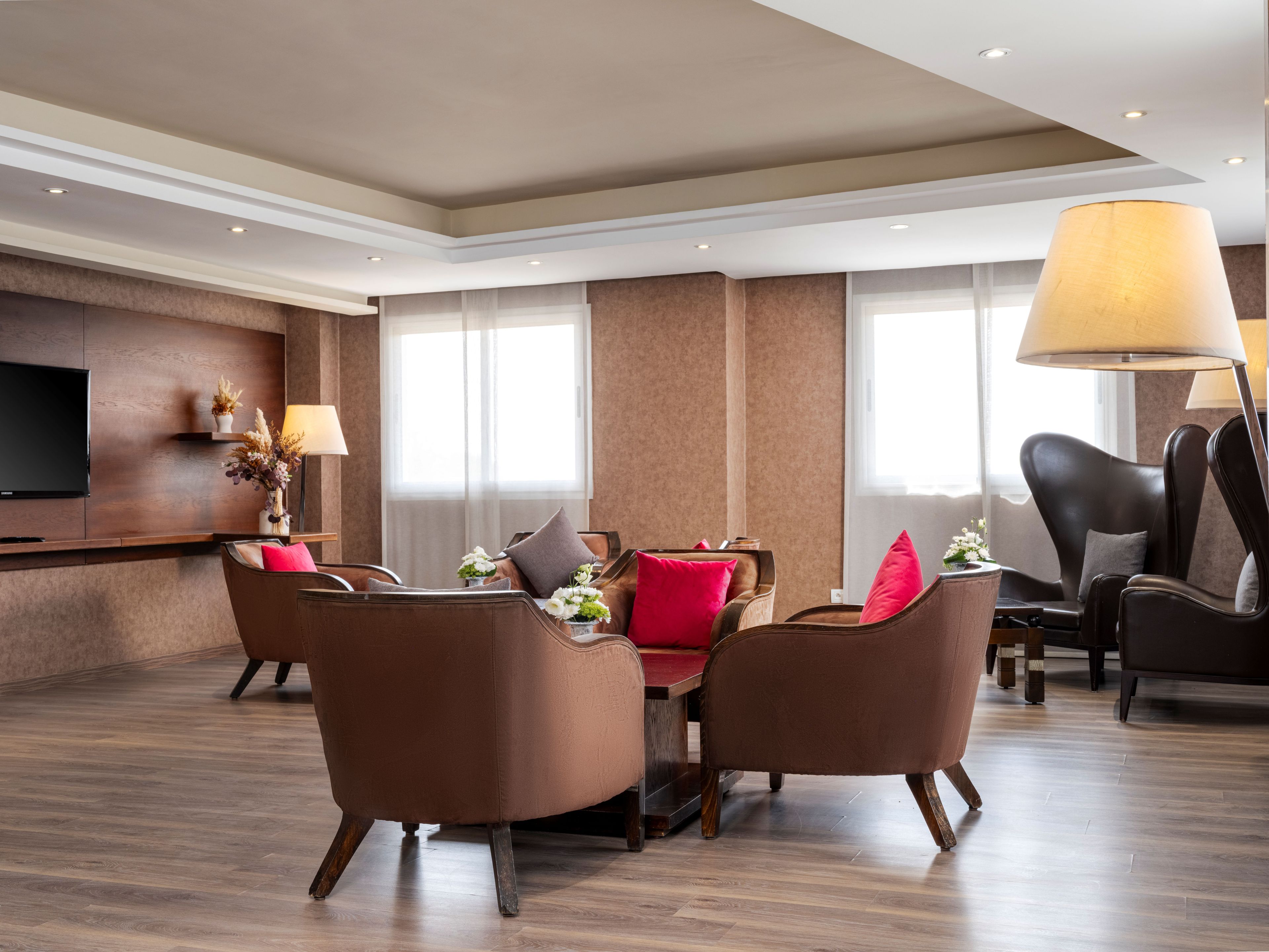 A modern hotel lobby with a flat-screen TV, seating area with sofas and armchairs, a large floor lamp, and a wooden reception desk.