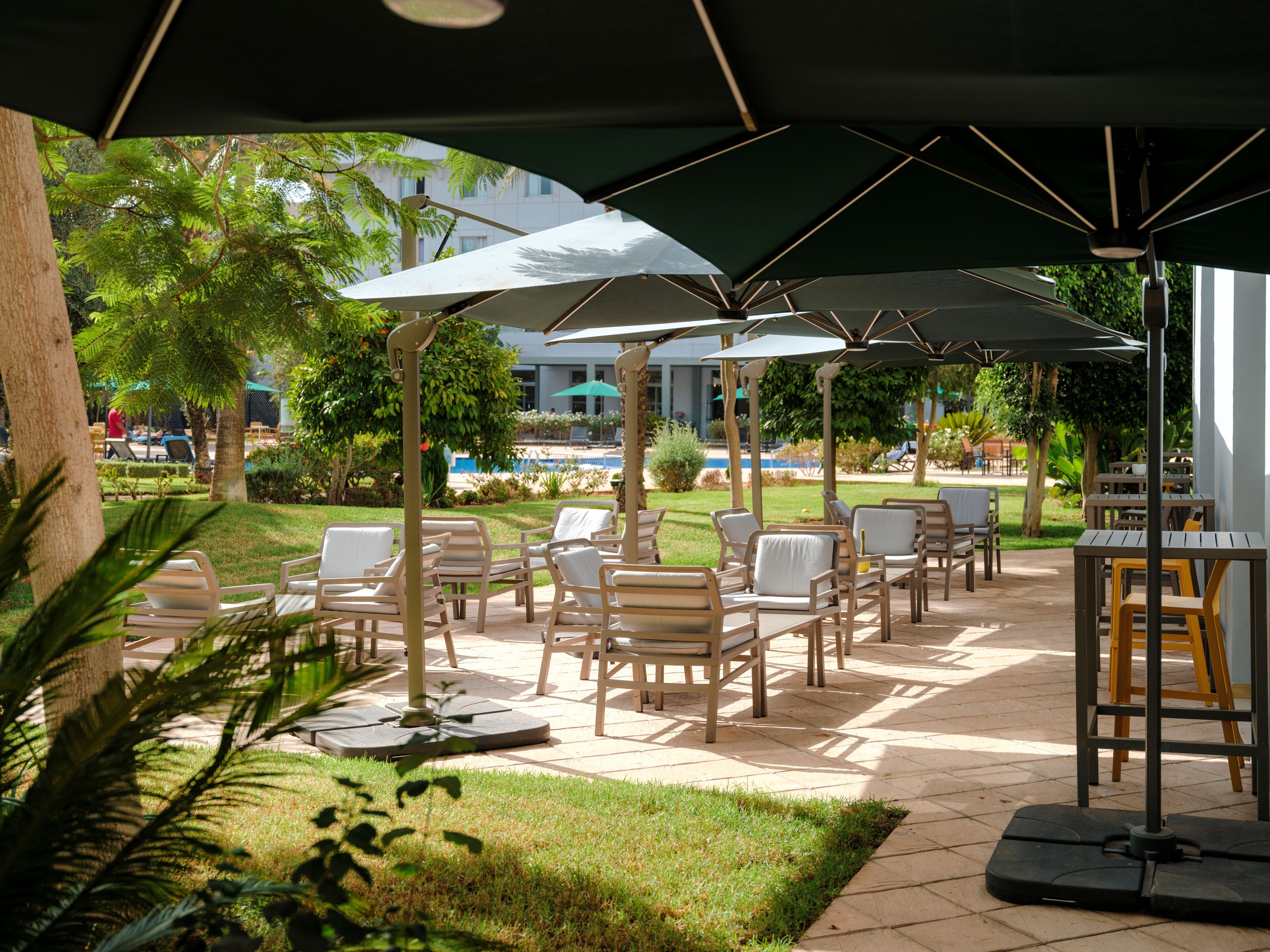 Un espace extérieur aménagé avec des parasols et des chaises, une piscine en arrière-plan et un bâtiment doté de fenêtres.