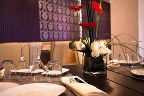 Una mesa de comedor dispuesta con platos blancos, copas de vino transparentes y un florero con flores blancas y rojas.