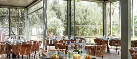 An outdoor dining area with wicker chairs and tables, surrounded by glass walls and large windows.