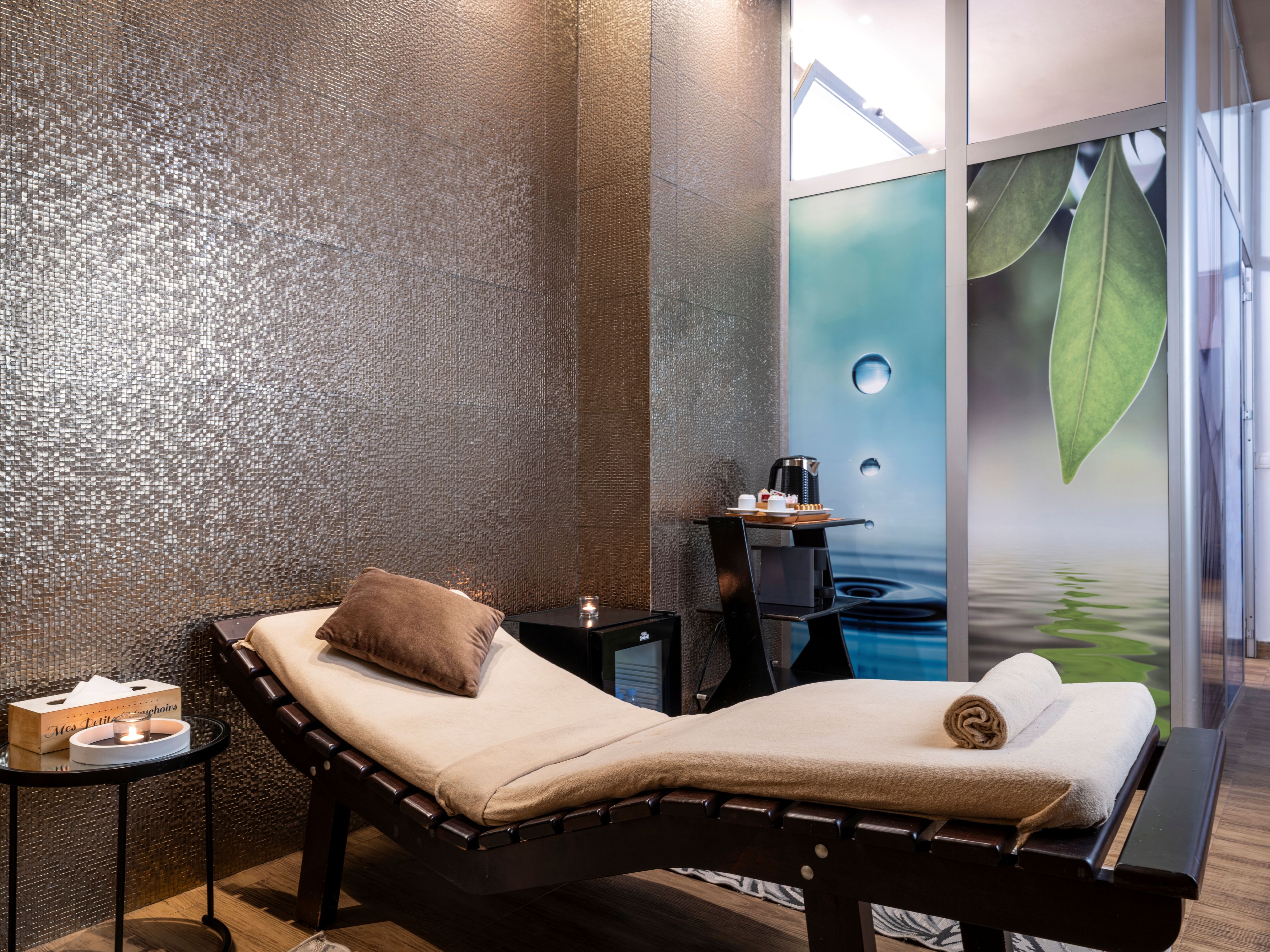 Une salle de spa équipée d’une table de massage, d’une petite table avec une bougie et d’un mur orné d’une fresque représentant des feuilles.