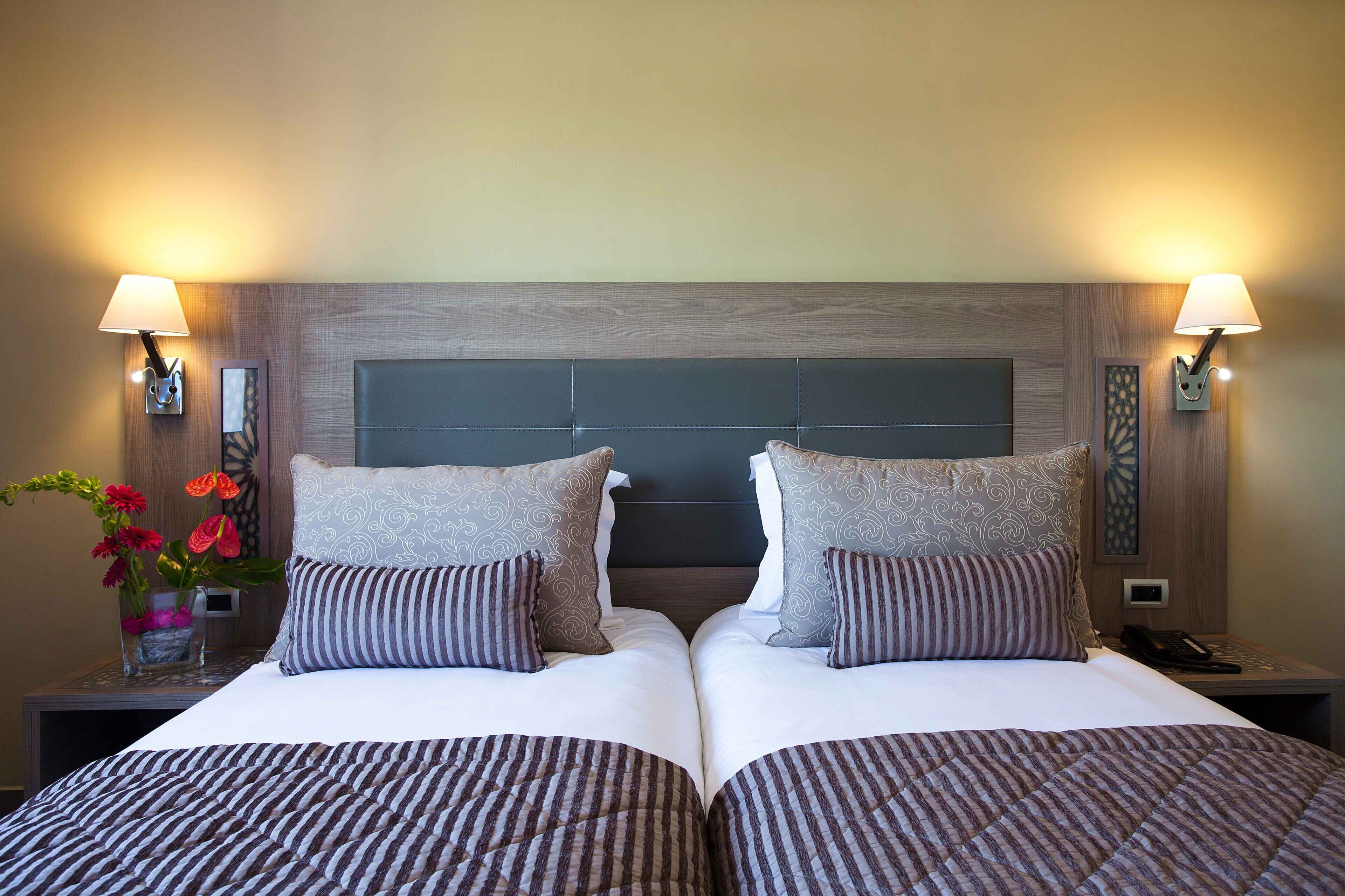 Una cama doble con ropa de cama a rayas, dos almohadas, mesitas de noche con lámparas y un jarrón con flores.