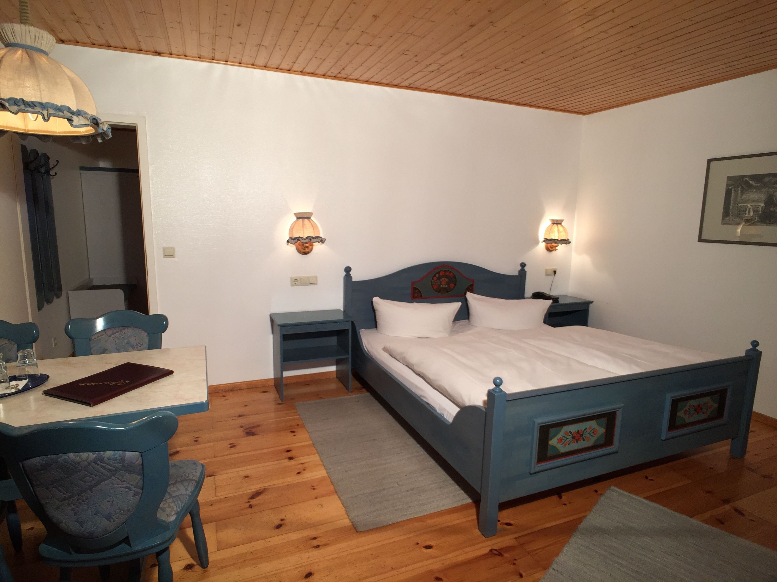 Un dormitorio con piso de madera, una cama con cabecera azul, dos mesitas de noche con lámparas, un escritorio con silla y un espejo.