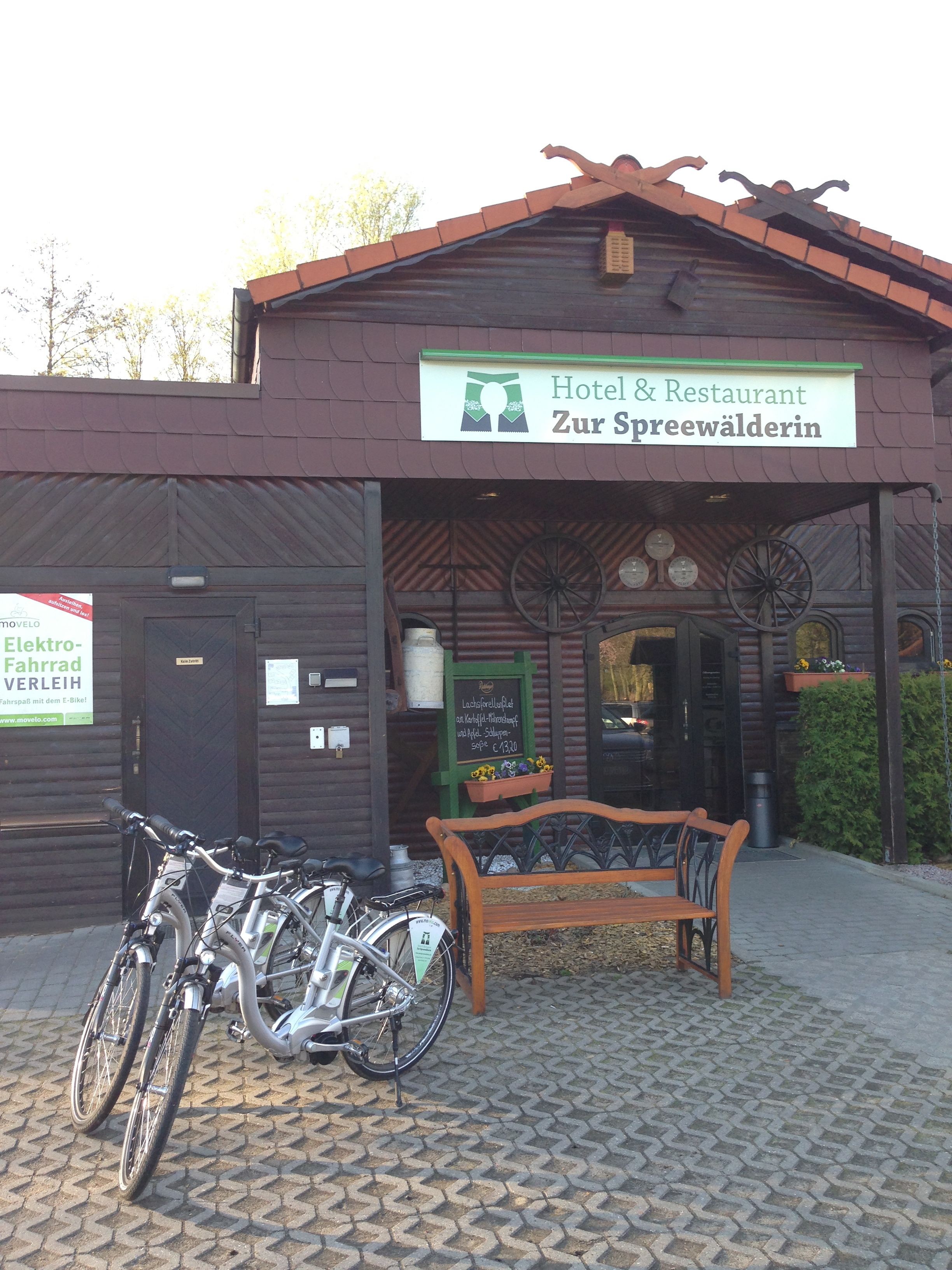 A building with a sign reading "Hotel & Restaurant Zur Spreewälderin." There are bicycles parked in front, a bench, and a wooden fence.