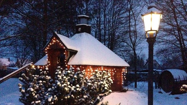 Une cabane en bois recouverte de neige, avec un lampadaire allumé, entourée d’arbres chargés de neige.