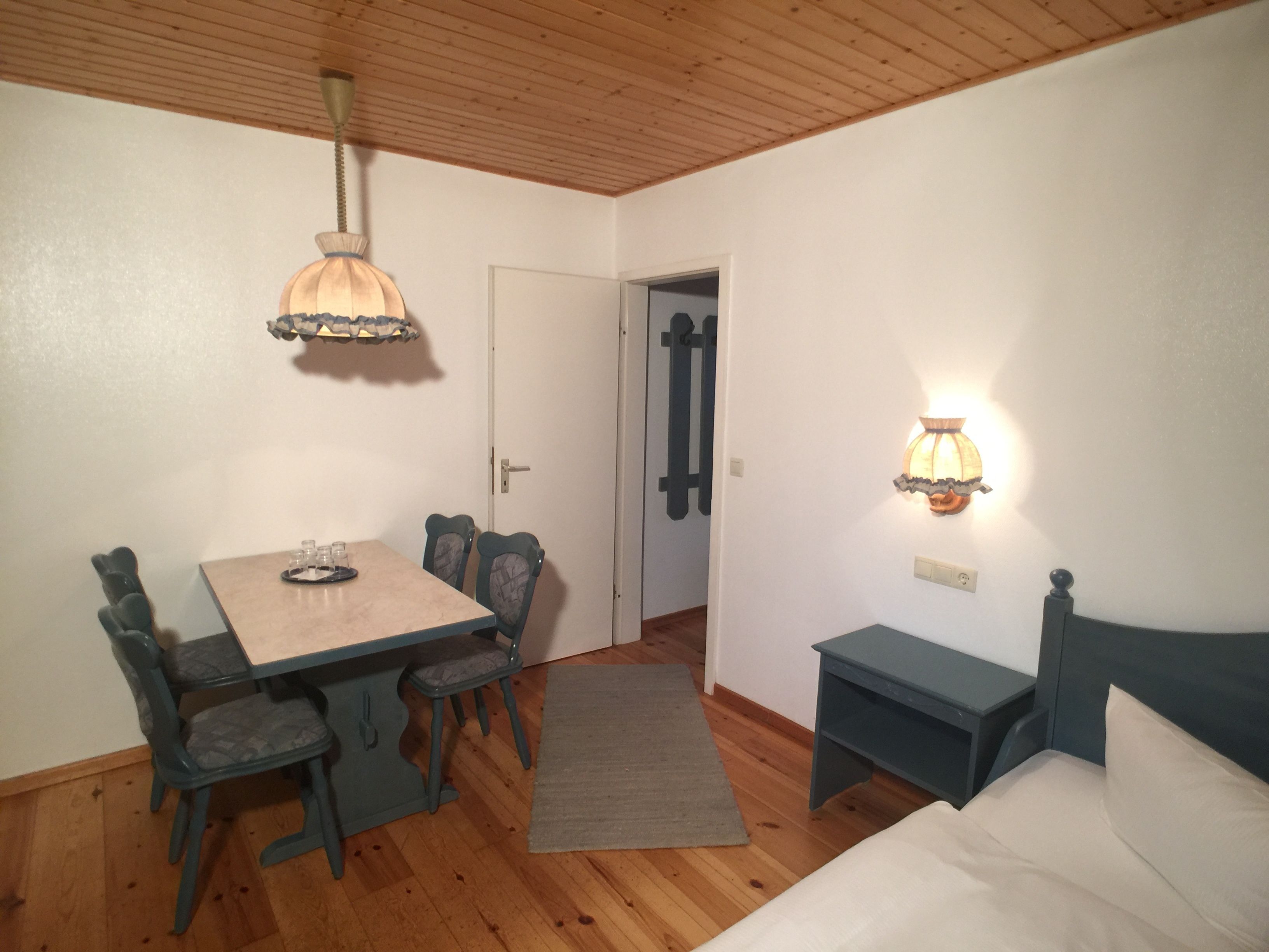Una stanza con soffitto in legno, un tavolo da pranzo con sedie, un letto e una lampada fissata al muro.